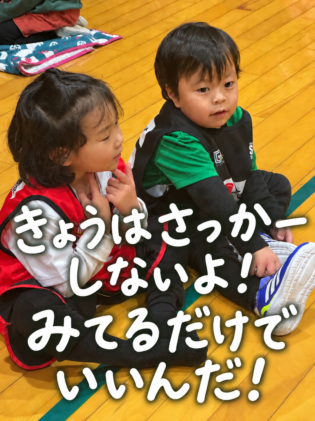 体育館で座って見学する2人の子どもたち。きょうはさっかーしないよ!! みてるだけでいいんだ!